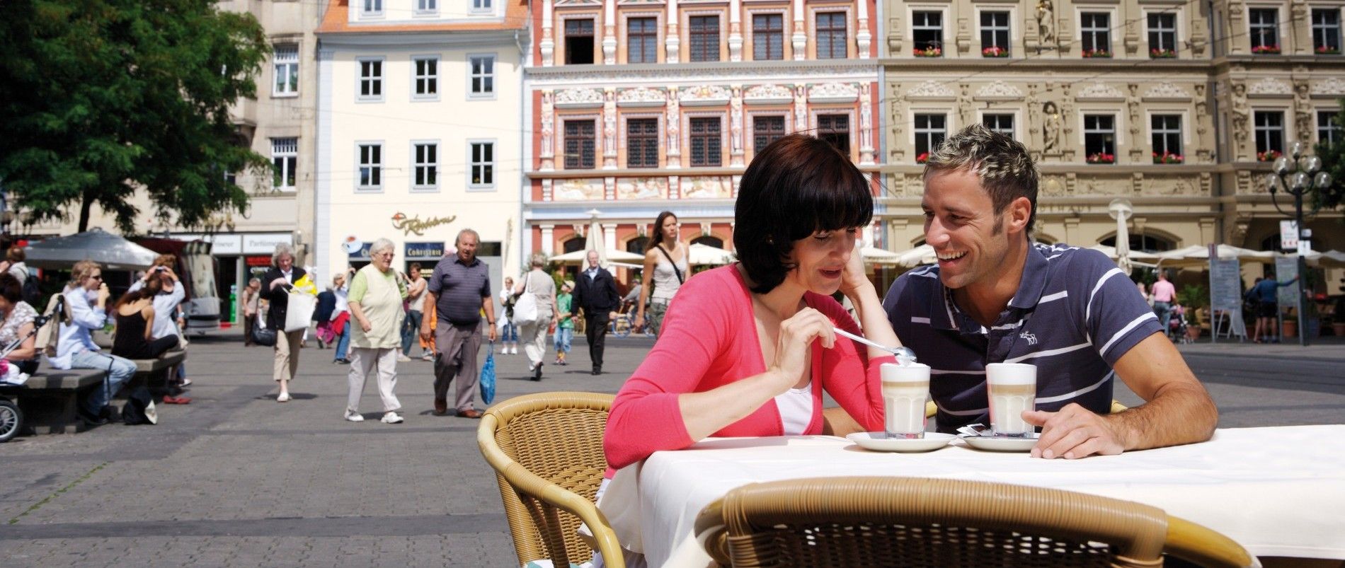 Ein lächelndes Paar genießt Kaffee an einem Tisch im Freien auf einem belebten Platz, umgeben von bunten historischen Gebäuden.