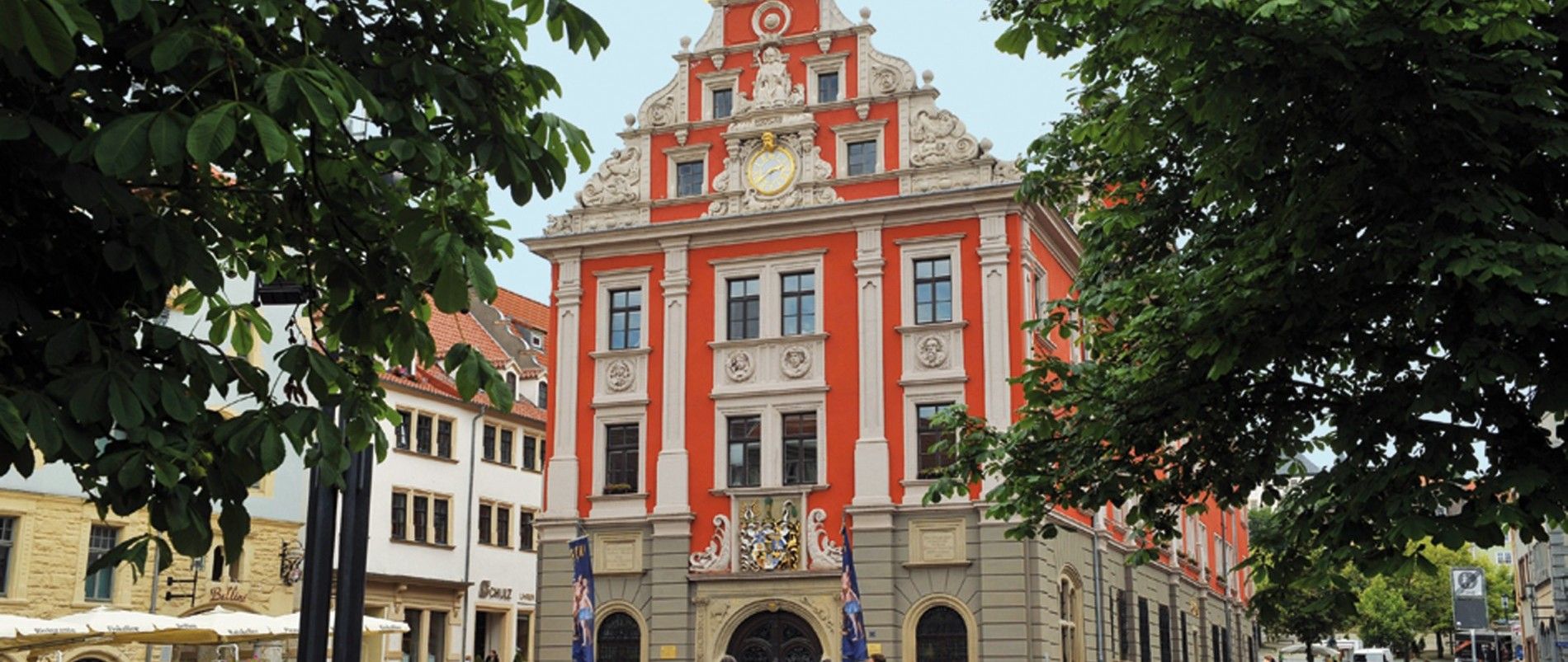 Historisches Gebäude mit rotem und beigefarbenem Fachwerkstil, umgeben von Bäumen.