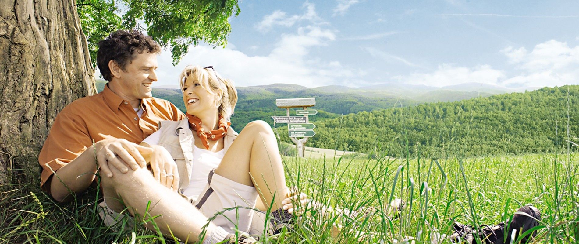 Paar sitzt lachend im Gras vor einem Baum mit einer grünen, hügeligen Landschaft im Hintergrund.