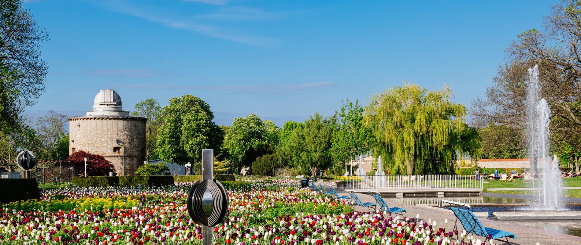 Ein gepflegter Park mit bunten Blumenbeeten, einem Brunnen und einem historischen runden Gebäude im Hintergrund.