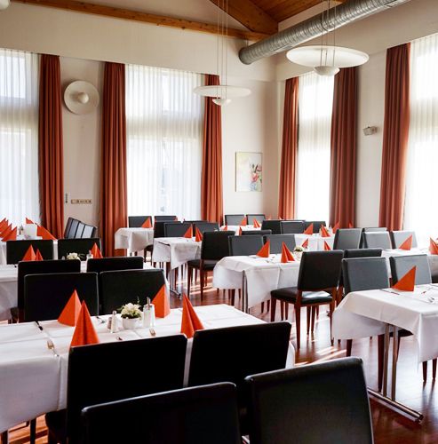 Elegantes Restaurant mit weißen Tischdecken, orange servierten Servietten und schwarzen Stühlen, umgeben von großen Fenstern mit orangefarbenen Vorhängen.