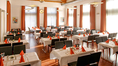 Elegantes Restaurant mit weißen Tischdecken, orange servierten Servietten und schwarzen Stühlen, umgeben von großen Fenstern mit orangefarbenen Vorhängen.