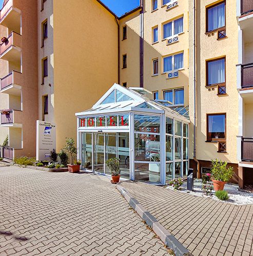 Eingang eines Hotels mit Glasüberdachung und beiger Fassade, umgeben von Pflanzen.