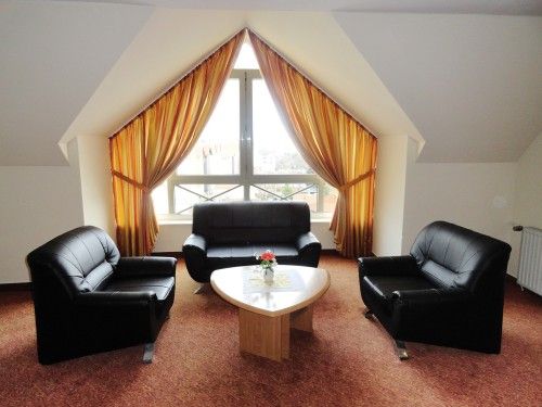 Geräumiger, eleganter Wohnbereich mit drei schwarzen Ledersesseln, einem Sofa und einem Holz-Couchtisch vor einem großen Fenster mit braunen Vorhängen.