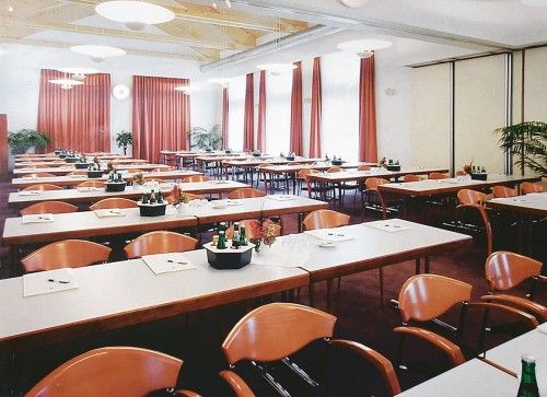 Konferenzraum mit mehreren Tischen und Stühlen, dekoriert mit Pflanzen und Vorhängen.