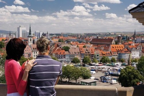 Ein Paar blickt auf eine Stadt mit roten Dächern und Kirchtürmen bei klarem Himmel.