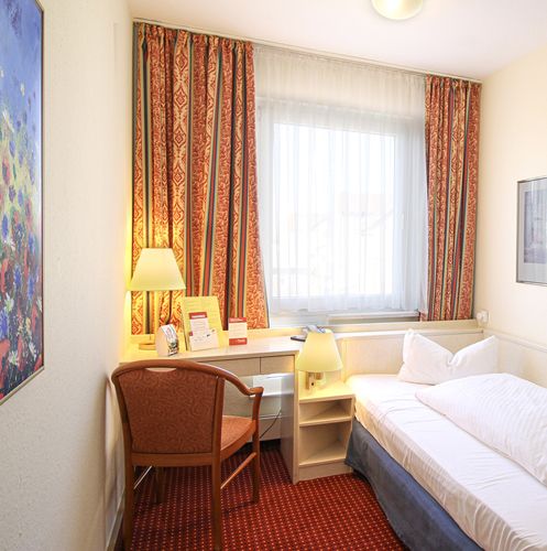 Gemütliches Hotelzimmer mit Einzelbett, Schreibtisch, Stuhl und bunten Vorhängen.