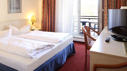 Helles Hotelzimmer mit Doppelbett, Schreibtisch, Fernseher und Balkon mit geöffneten Vorhängen.