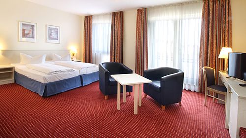 Helles Hotelzimmer mit zwei Einzelbetten, zwei Sesseln, einem Tisch und roten Teppichboden.