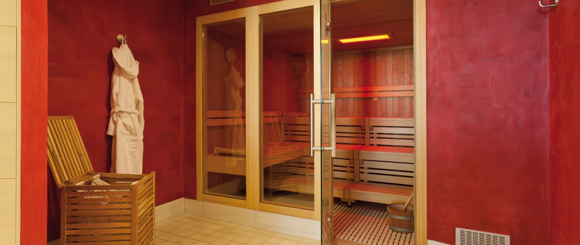 Sauna mit Holzbänken, Glastüren und roter Wand, ein Holzkorb mit Bademantel daneben.